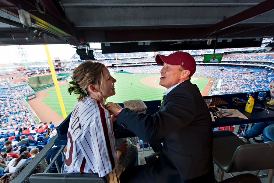 A wedding couple having a food fight with popcorn and laughing at a Phillies game.